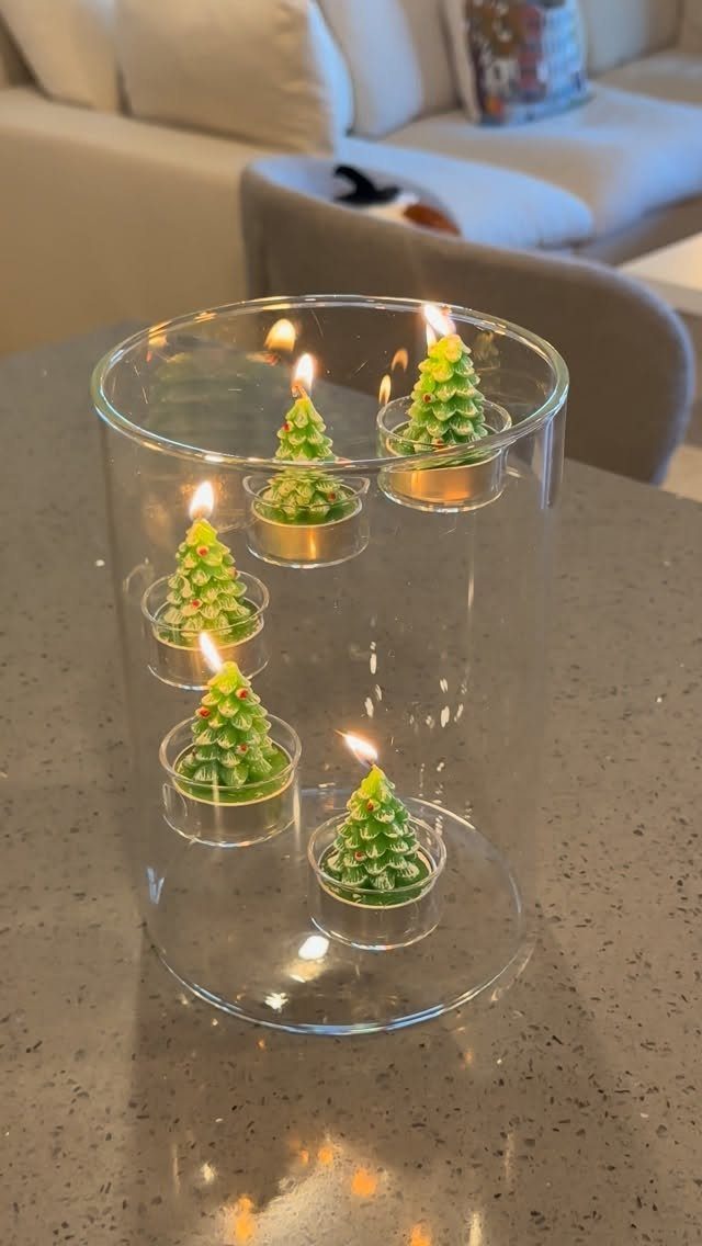 Green tree-shaped candles lit inside a clear glass cylinder on a grey counter.