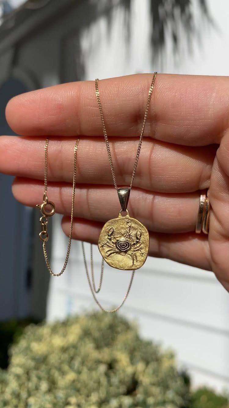 Gold necklace with a crab pendant held in a person's hand outdoors.