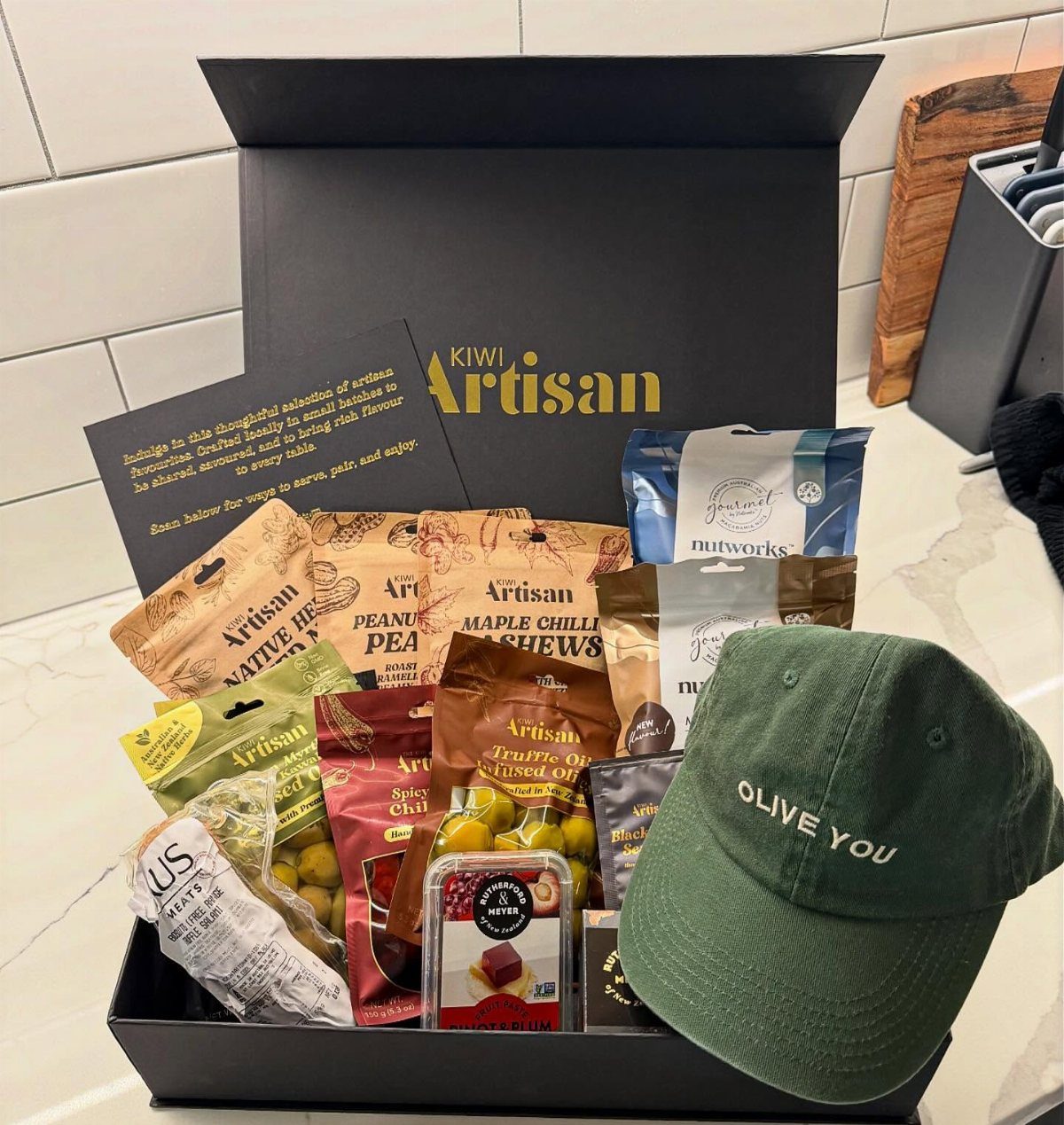 Artisan snack box filled with gourmet treats and a green olive-themed baseball cap.