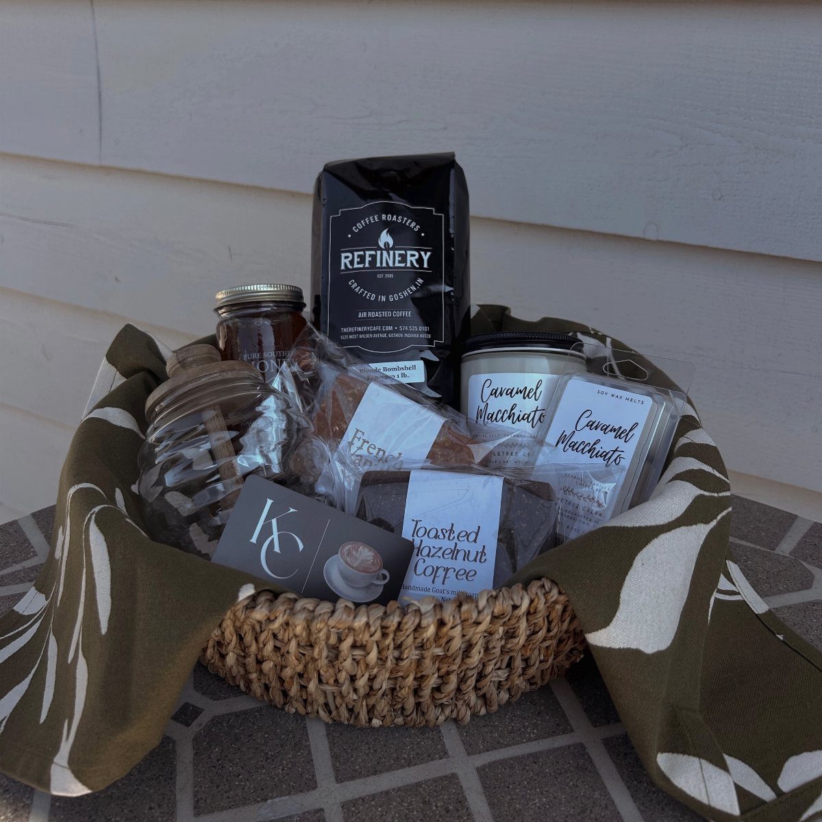 Woven gift basket containing coffee beans, flavored coffee jars, and treats in fabric.