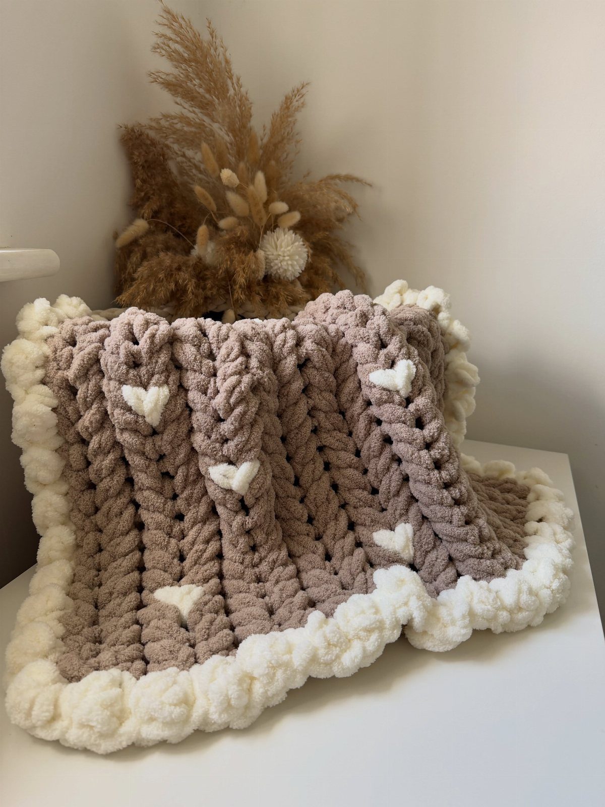 Chunky knit mauve blanket with white hearts and cream border on white surface.