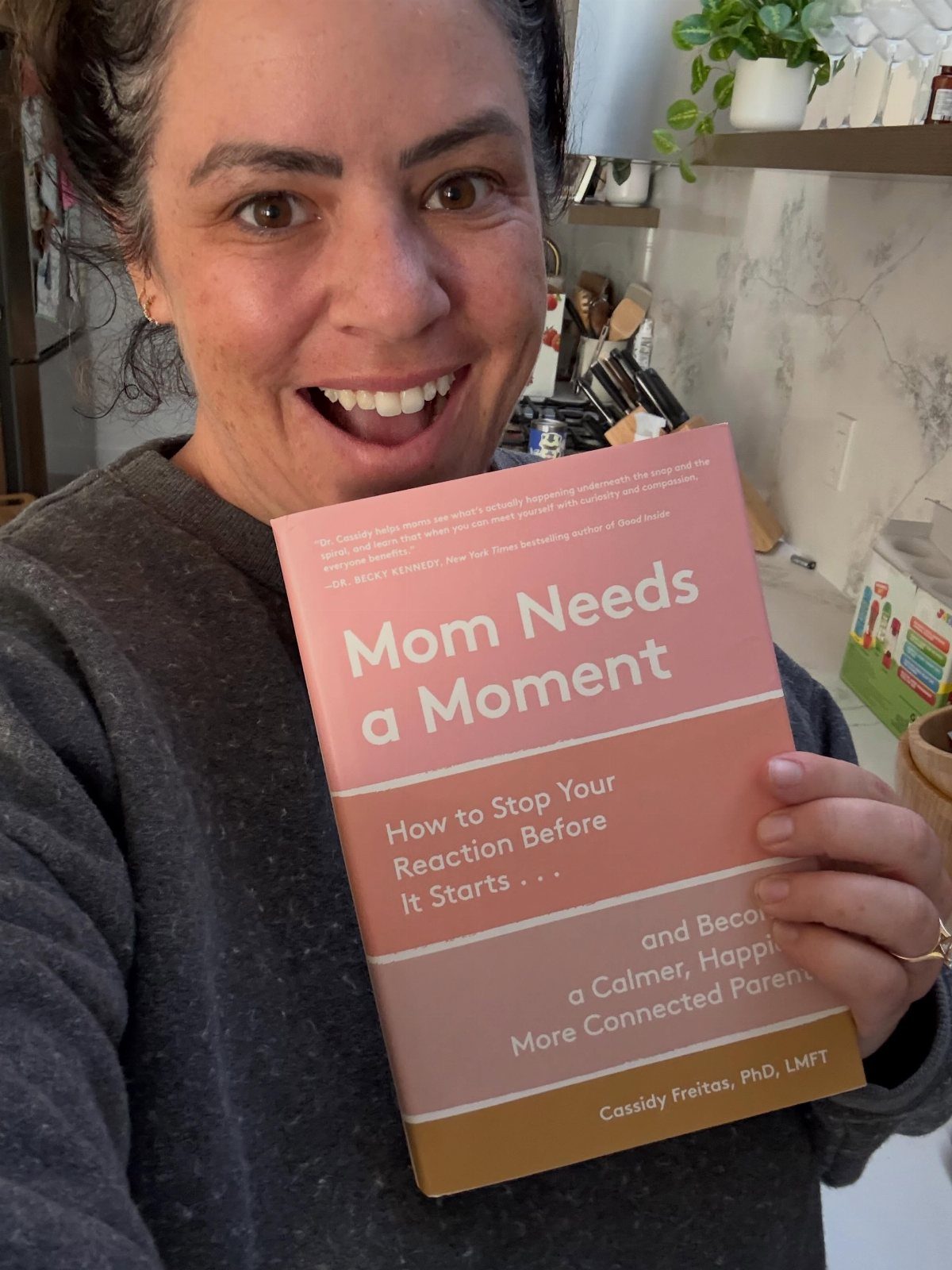 Smiling woman holding a parenting book titled Mom Needs a Moment.