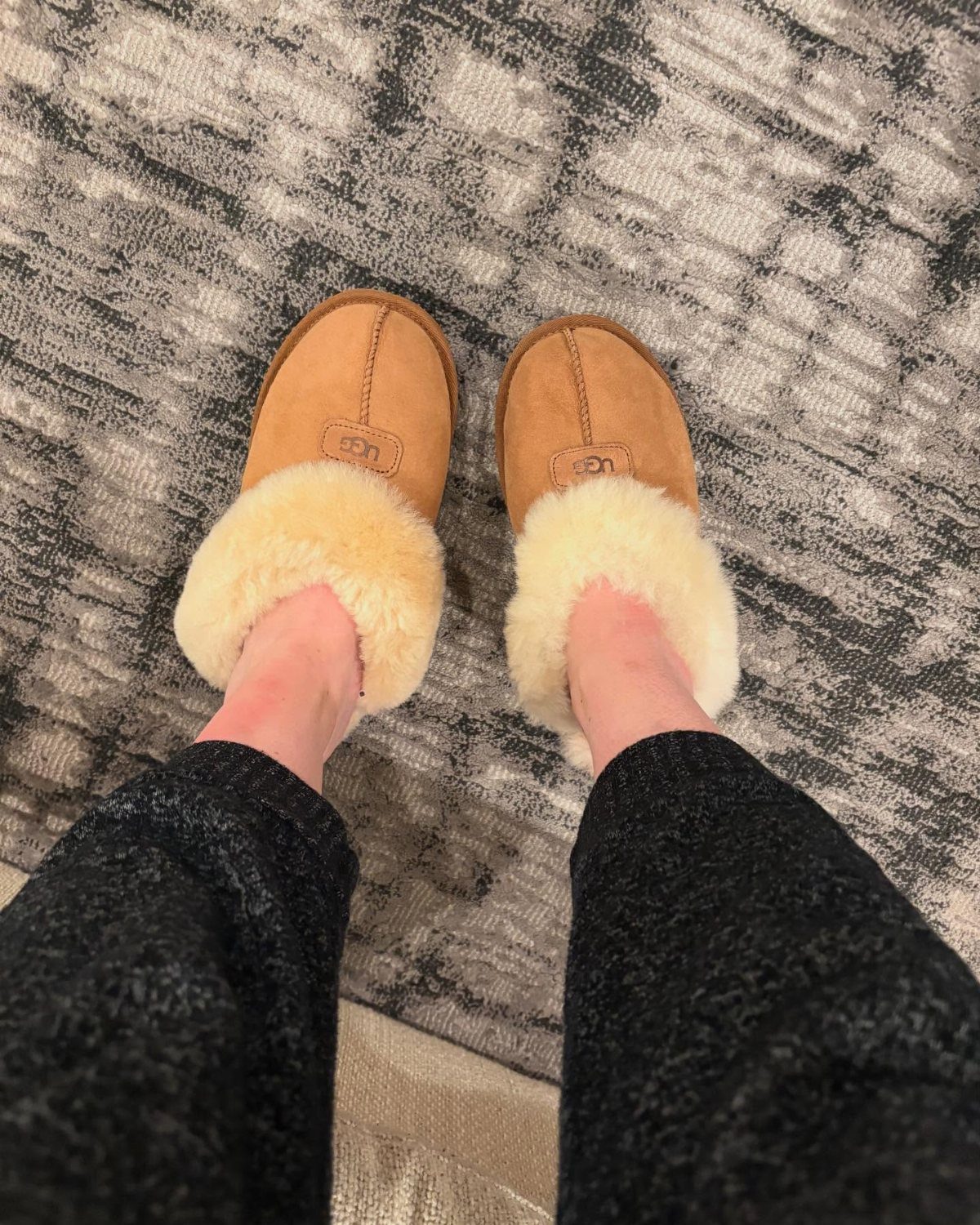 Tan suede slippers with white fluffy lining worn on a grey patterned rug.