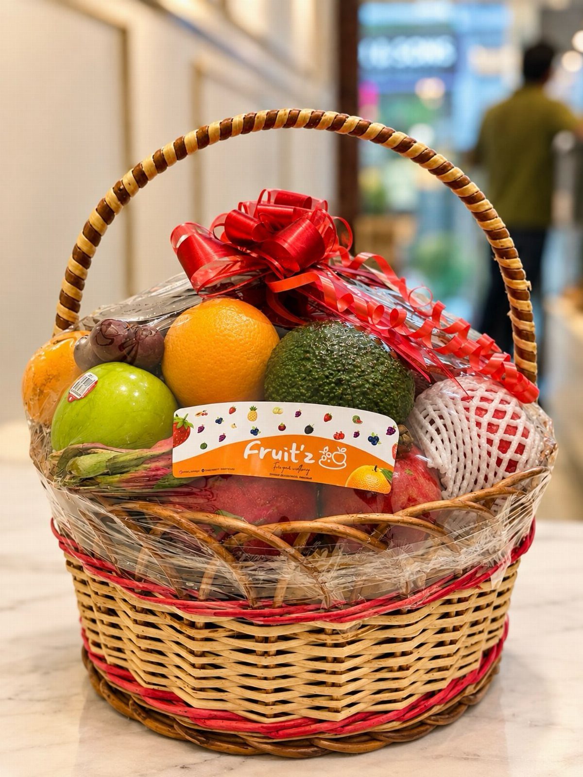 Woven fruit basket containing a green apple, orange, and avocado with red bow.