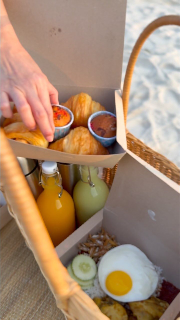 Woven picnic basket with croissants, juices, and breakfast food on a sandy beach.