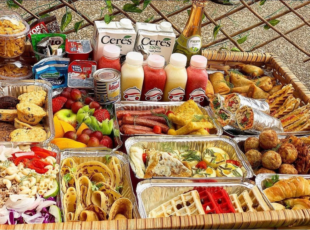 Woven picnic basket filled with waffles, croissants, fruit, and bottled drinks outdoors.