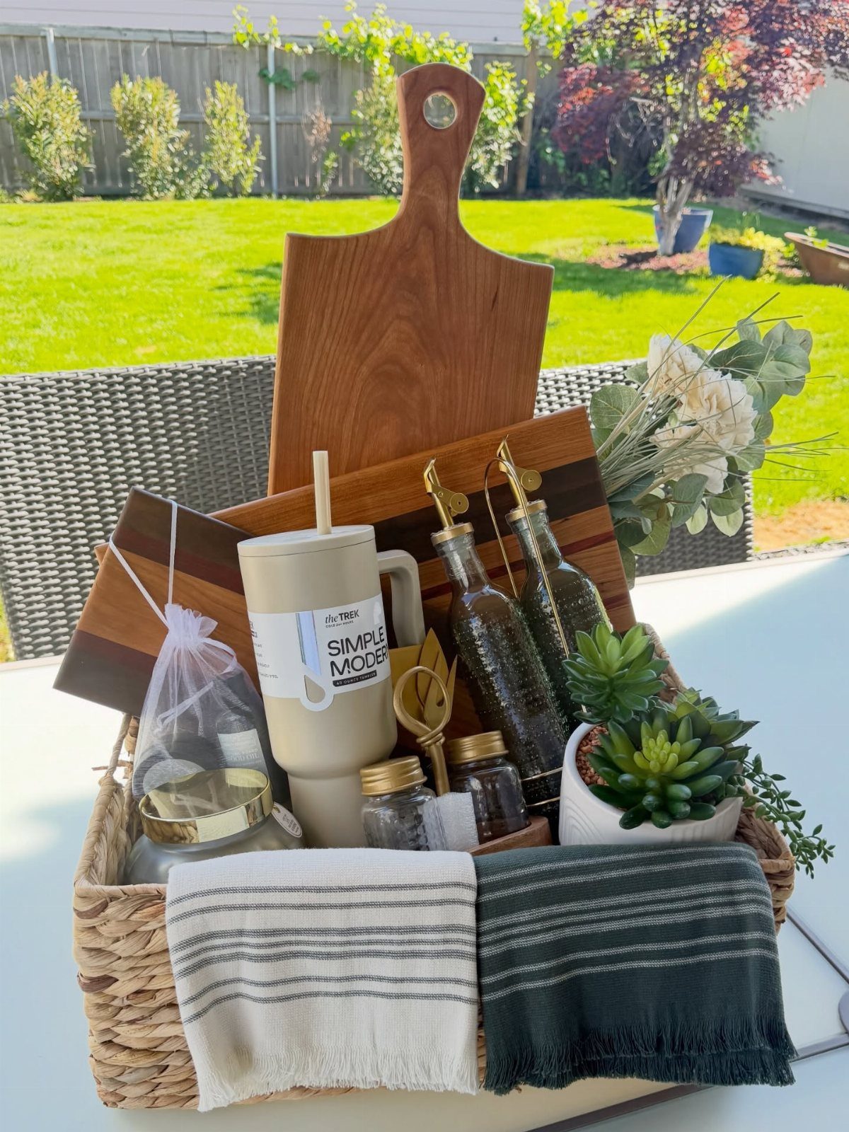 Woven gift basket containing a wooden board, mug, bottles, and striped towels outdoors.