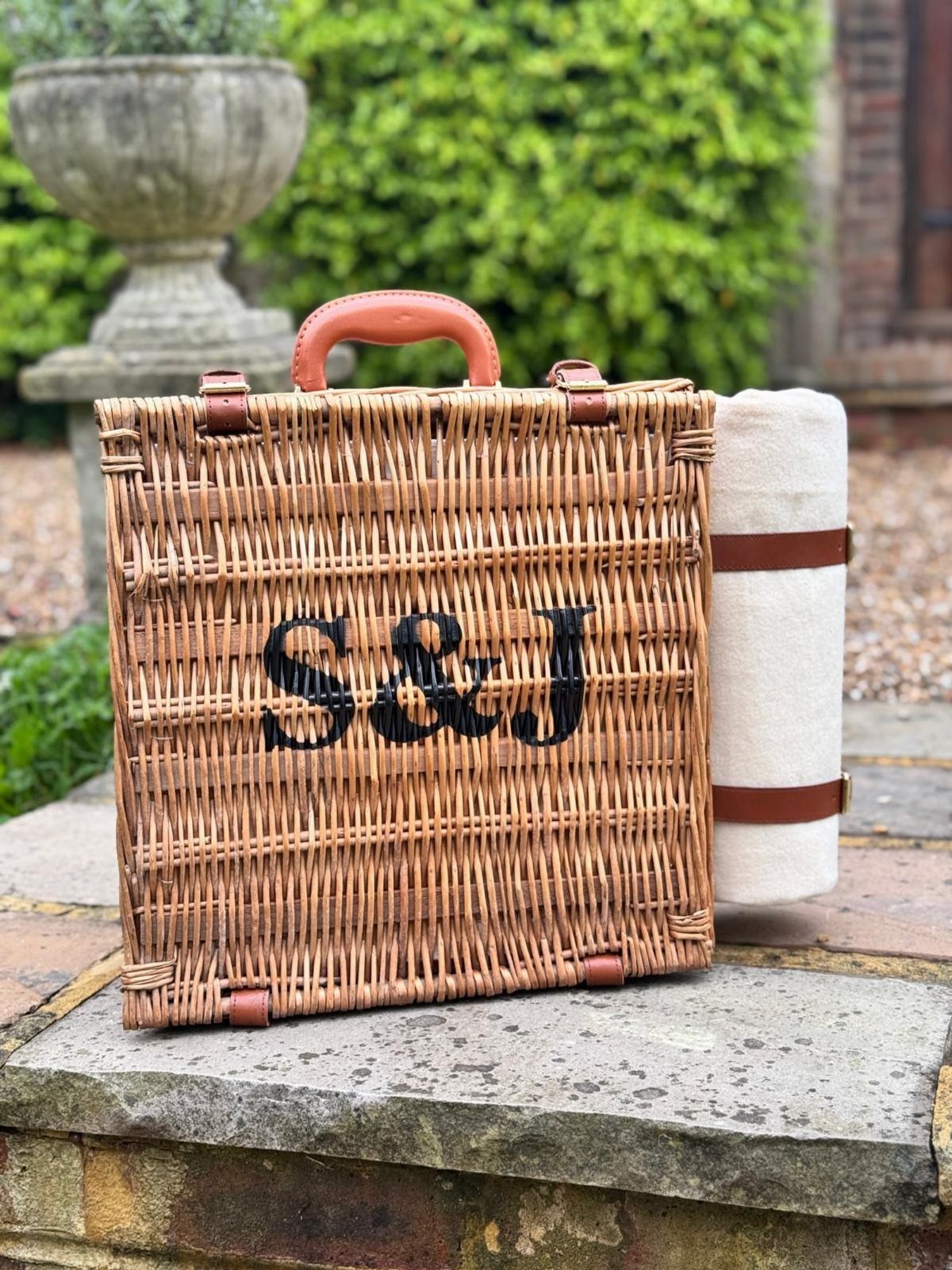 Woven wicker picnic basket with S&J initials and a rolled white blanket.