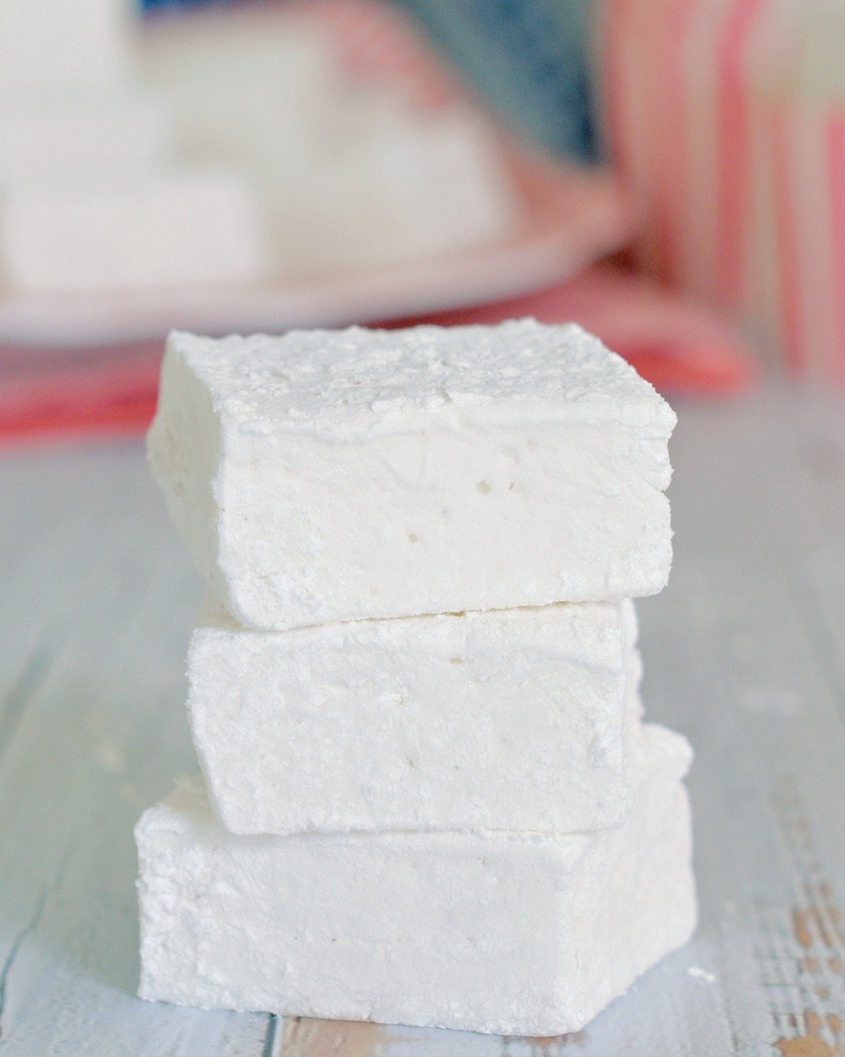 Three square white homemade marshmallows stacked on a light wooden surface.