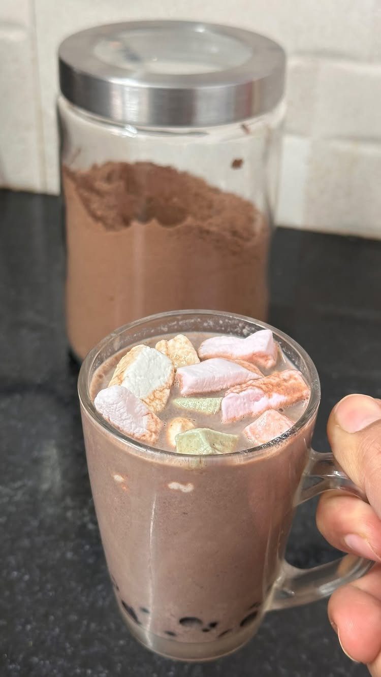Hot chocolate with colorful marshmallows in a mug next to a cocoa powder jar.