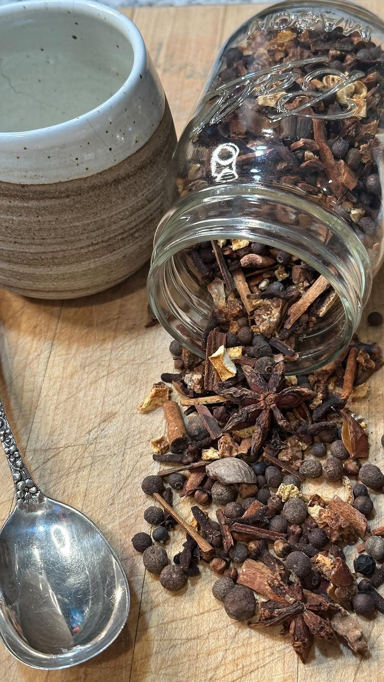 Mulling spices spilling from a glass jar onto a wooden board with a spoon.