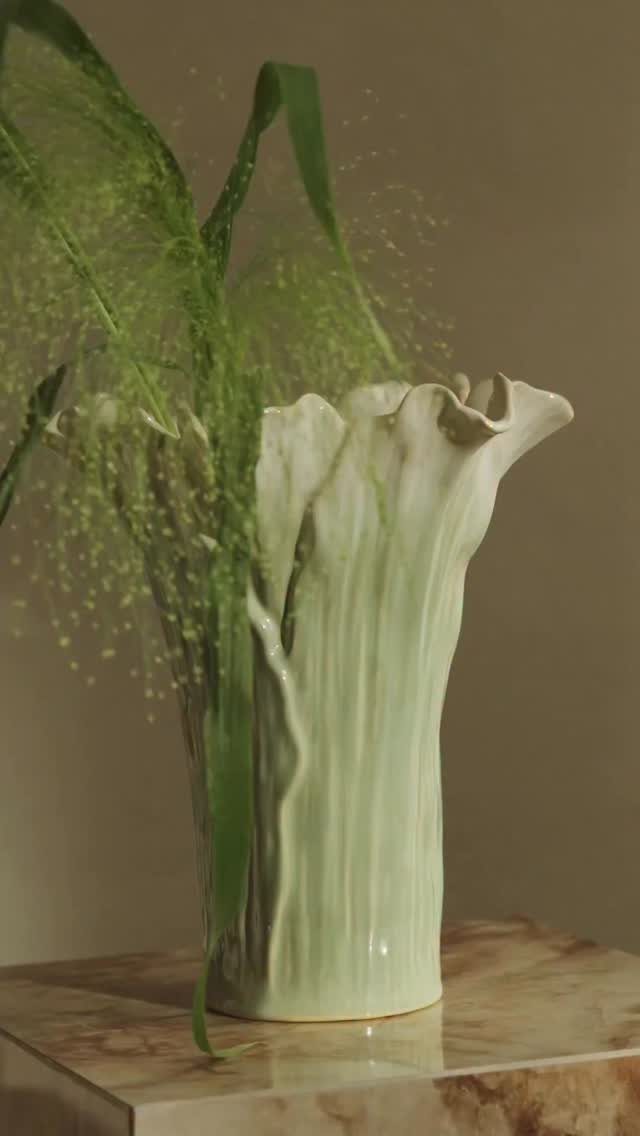 Organic light green ceramic vase holding green foliage on a marble surface.