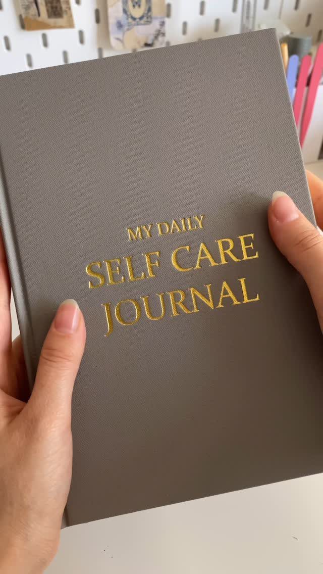 Grey hardcover self care journal with gold lettering held by two hands.