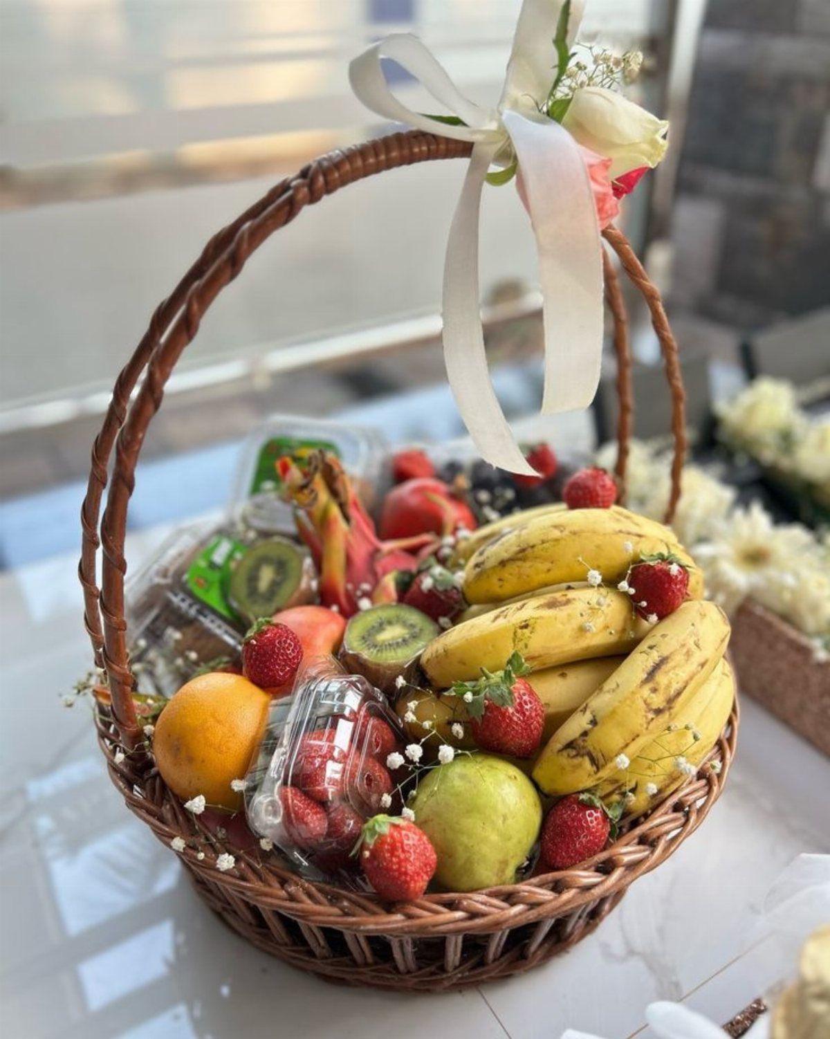 Wicker gift basket filled with bananas, strawberries, kiwi, and apples with white ribbon.