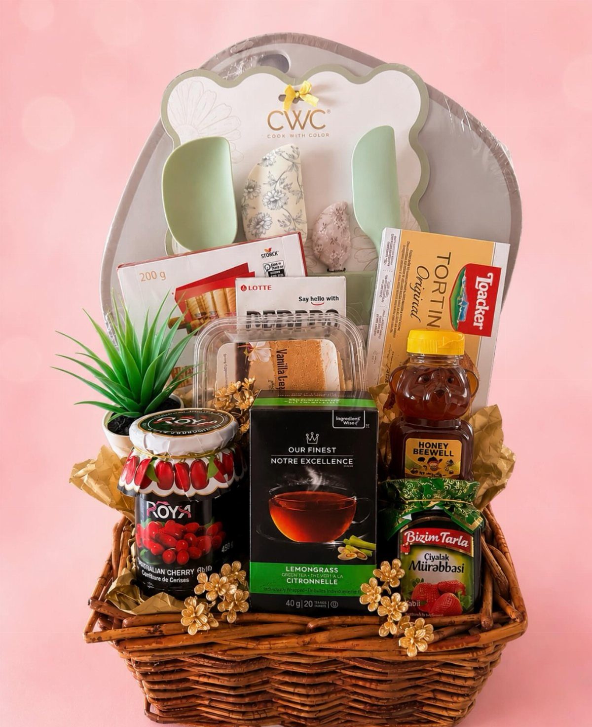 Wicker gift basket containing honey, jam, tea, and cookies on pink background