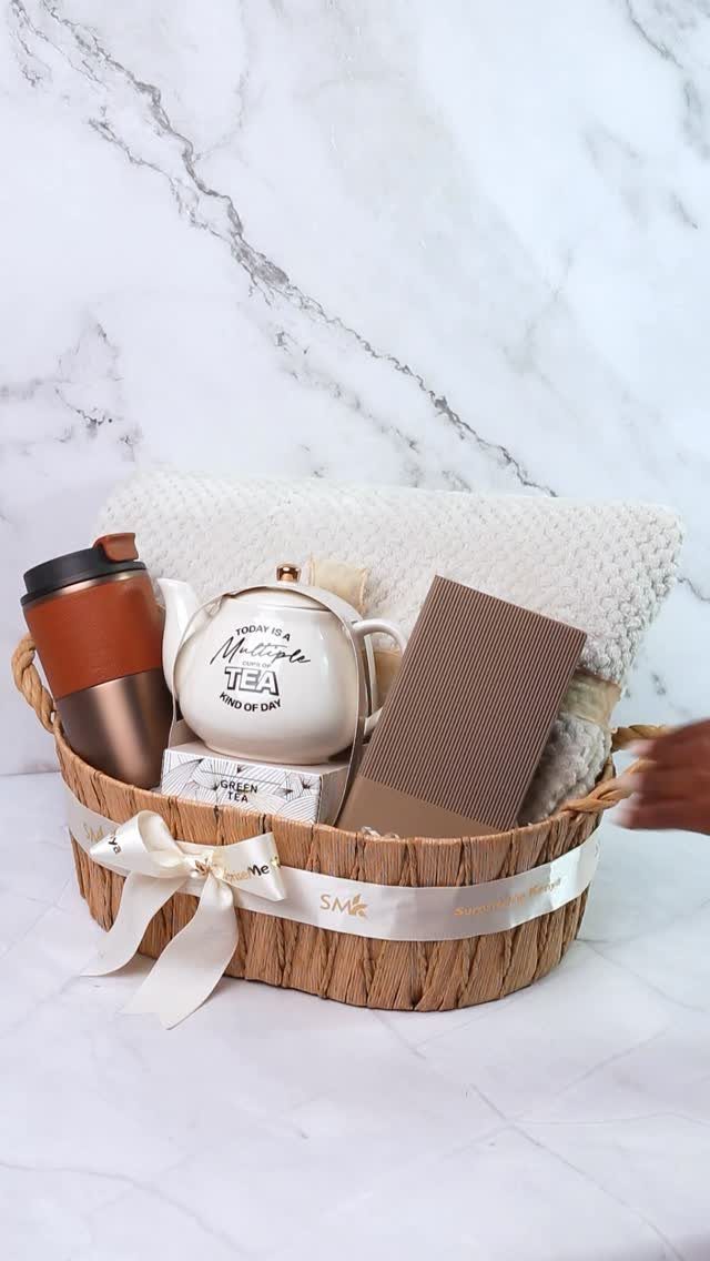 Woven gift basket with a teapot, travel mug, notebook, and white blanket.