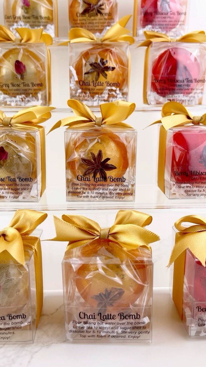 Tea bombs in clear gift boxes with gold ribbons and flavor labels.
