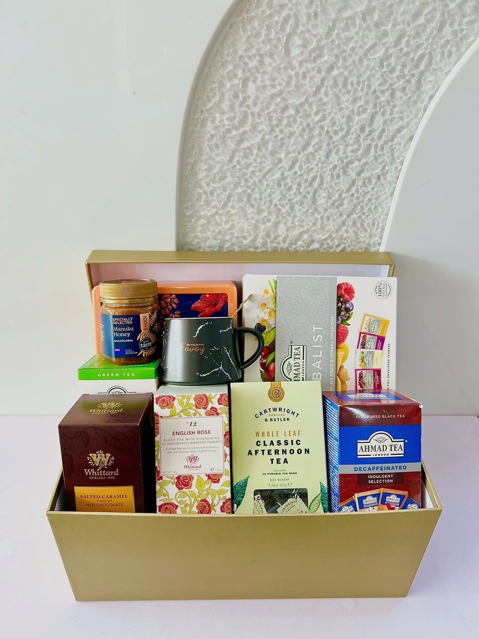 Gold gift box containing assorted tea boxes, a black mug, honey, and sweets.