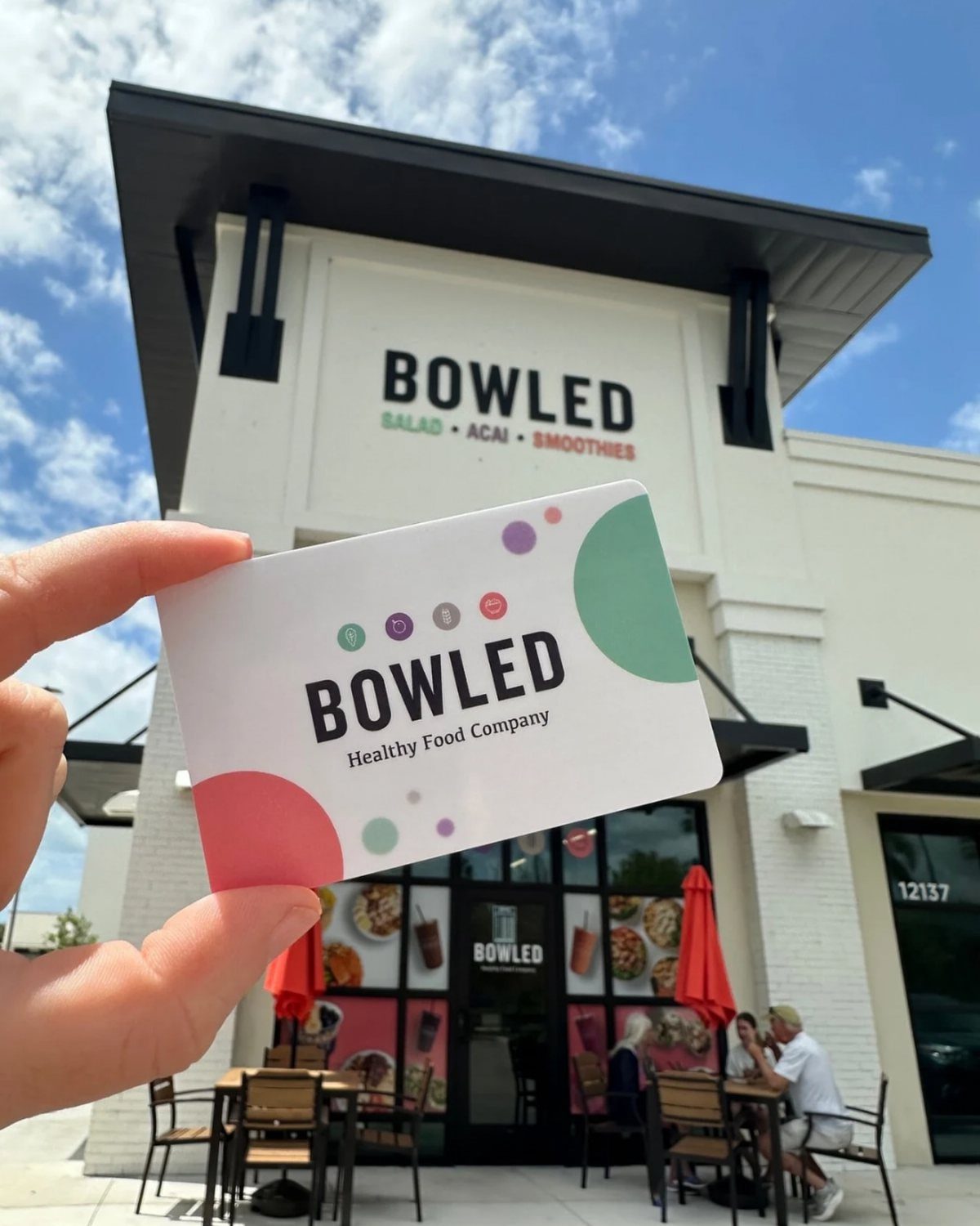 8 Teacher Gifts They Actually Want, From Notes to Totes 7 Gift card for Bowled Healthy Food Company held in front of the restaurant building.