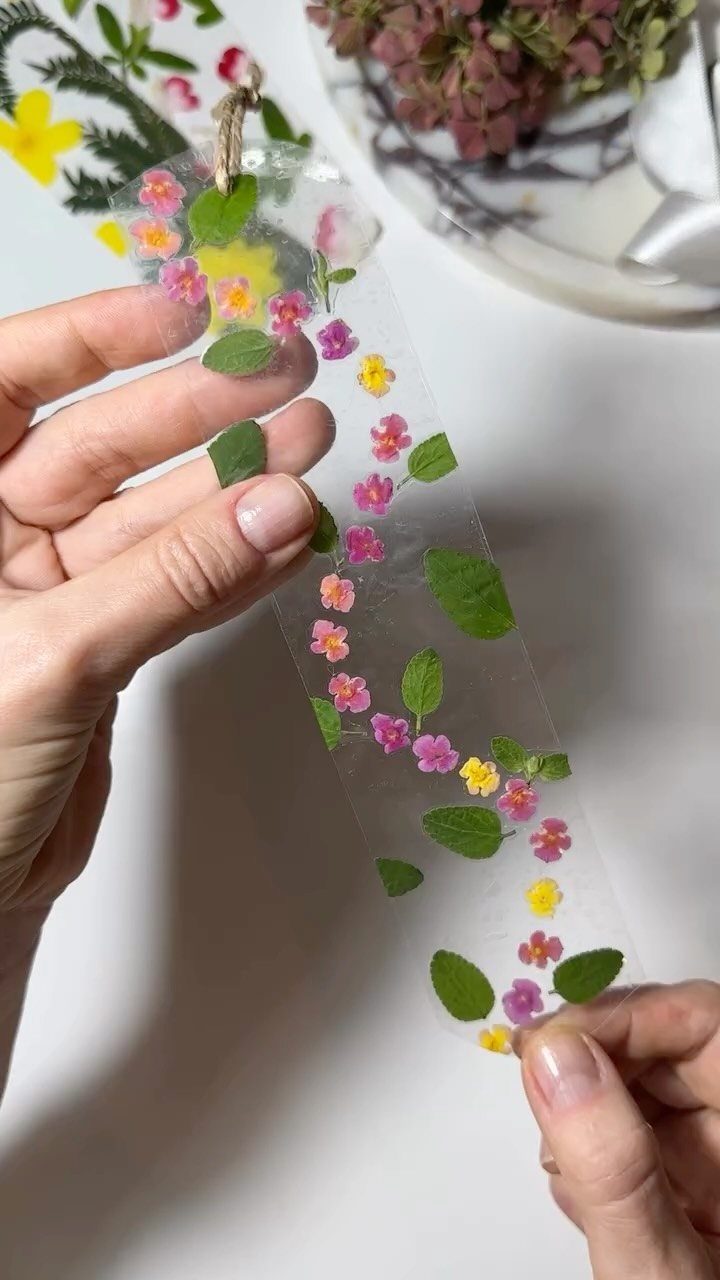 12 Useful Homemade Christmas Gifts People Will Use 12 Transparent bookmark with pressed colorful flowers and green leaves held by two hands.