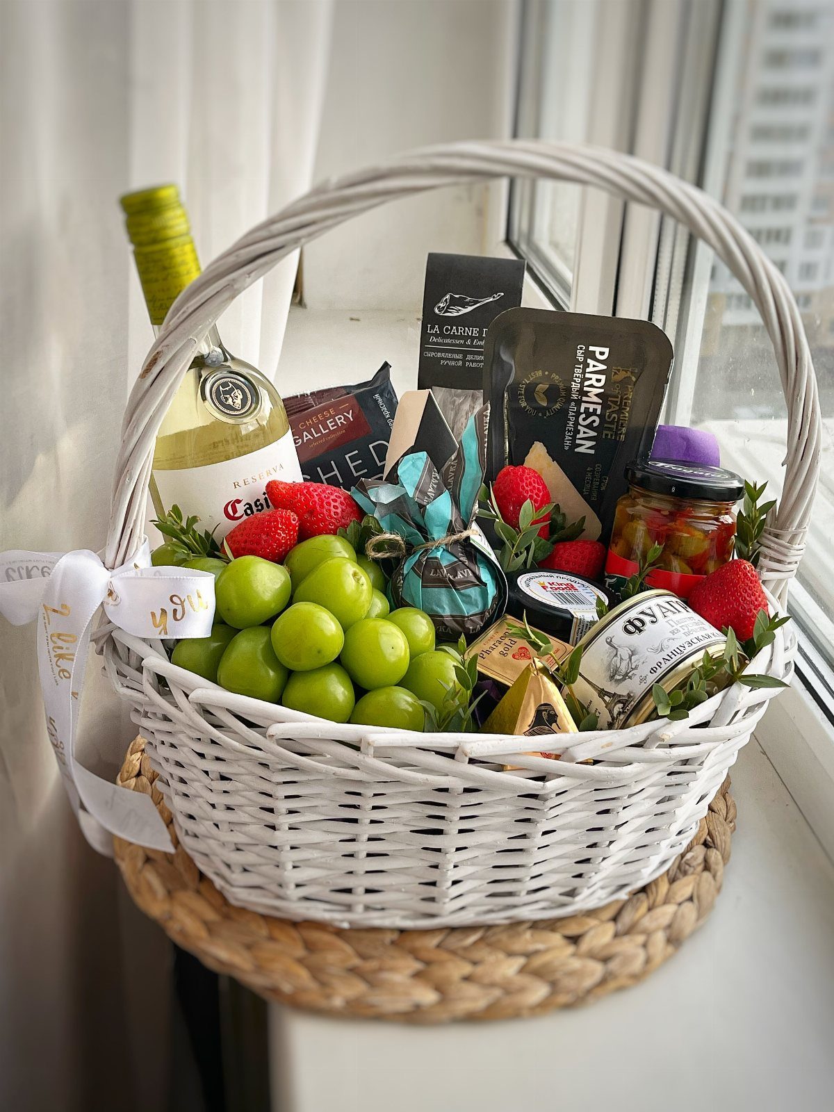 White wicker gift basket with white wine, green grapes, strawberries, and gourmet cheeses.